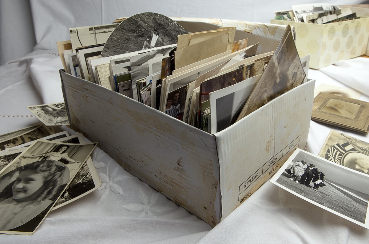 A shoebox full of old photographs, surrounded by even more photos, sits on a white tablecloth.