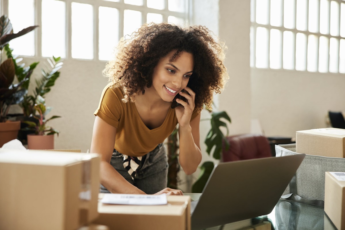A small business owner surrounded by shipping boxes answers a call while looking up a document on her laptop.