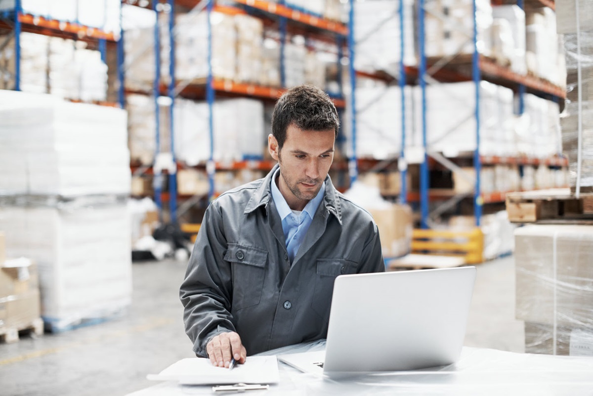 A man taking notes while looking at a laptop screen and standing in a stocked warehouse.