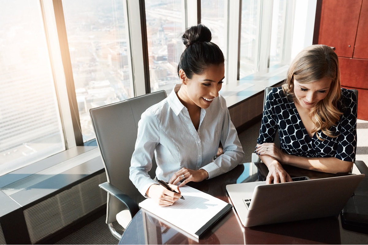 Two women sit at a desk in an office, looking at a laptop. One of the women is writing on a notepad.