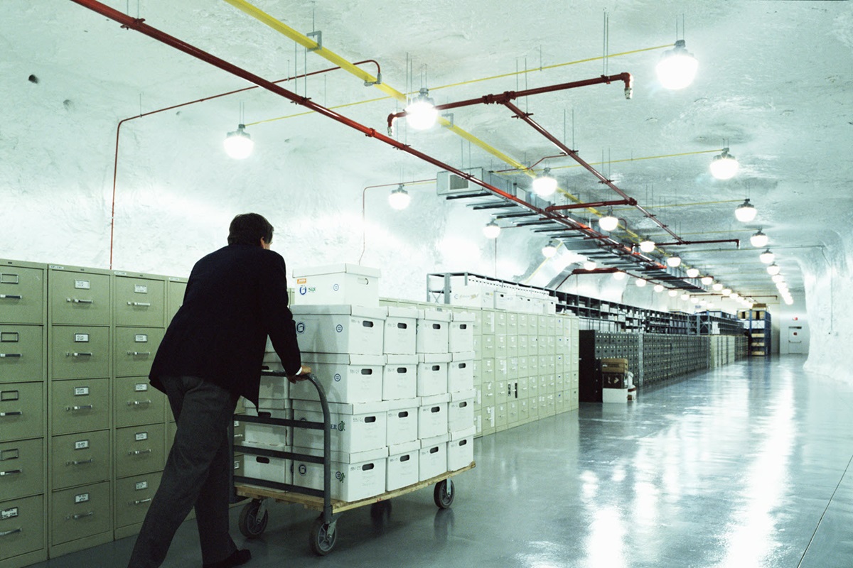 An office worker pushes a cart loaded with file boxes down a wide hallway lined with file cabinets.