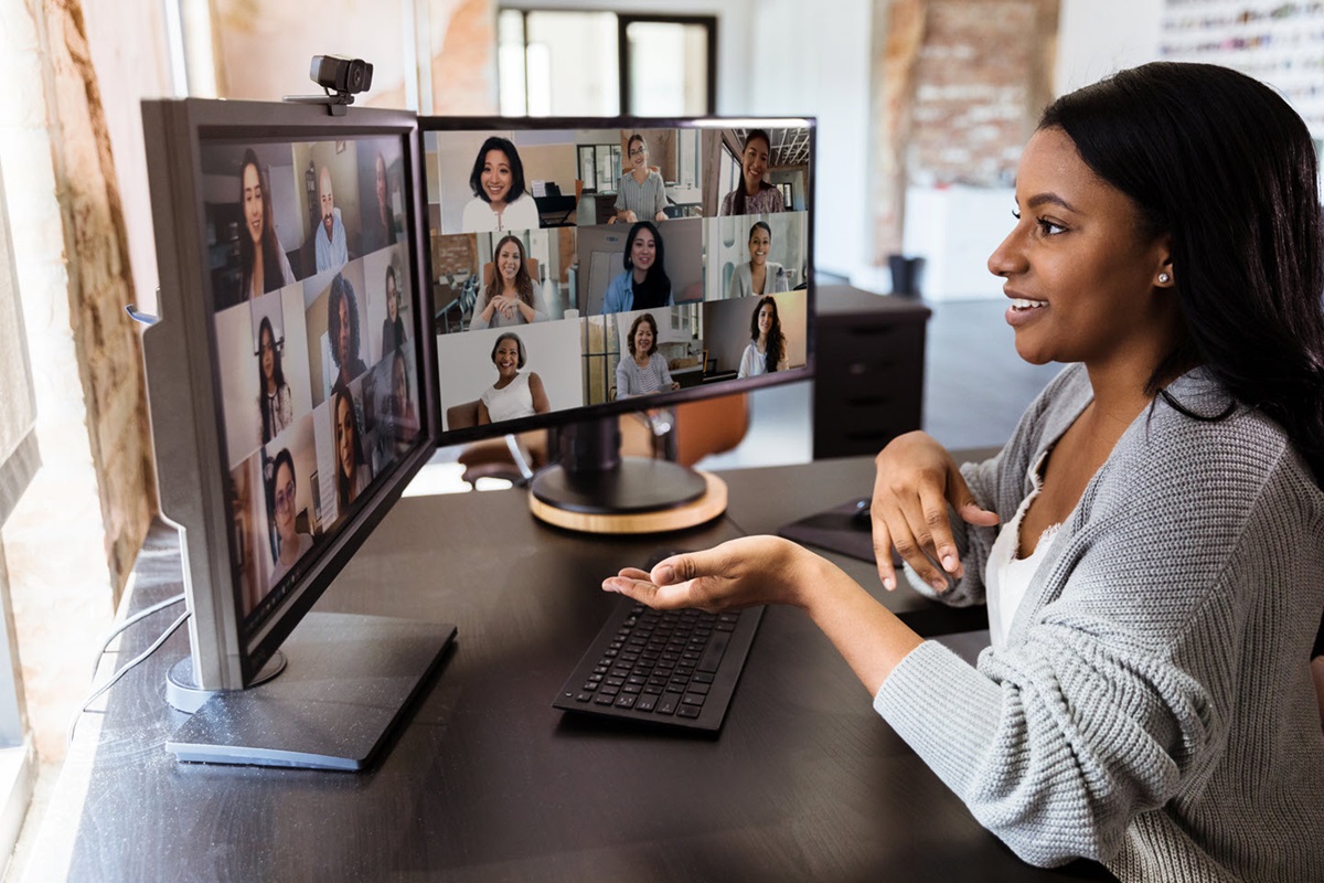 A woman in a grey sweater sits at a desk while participating in a group video call.