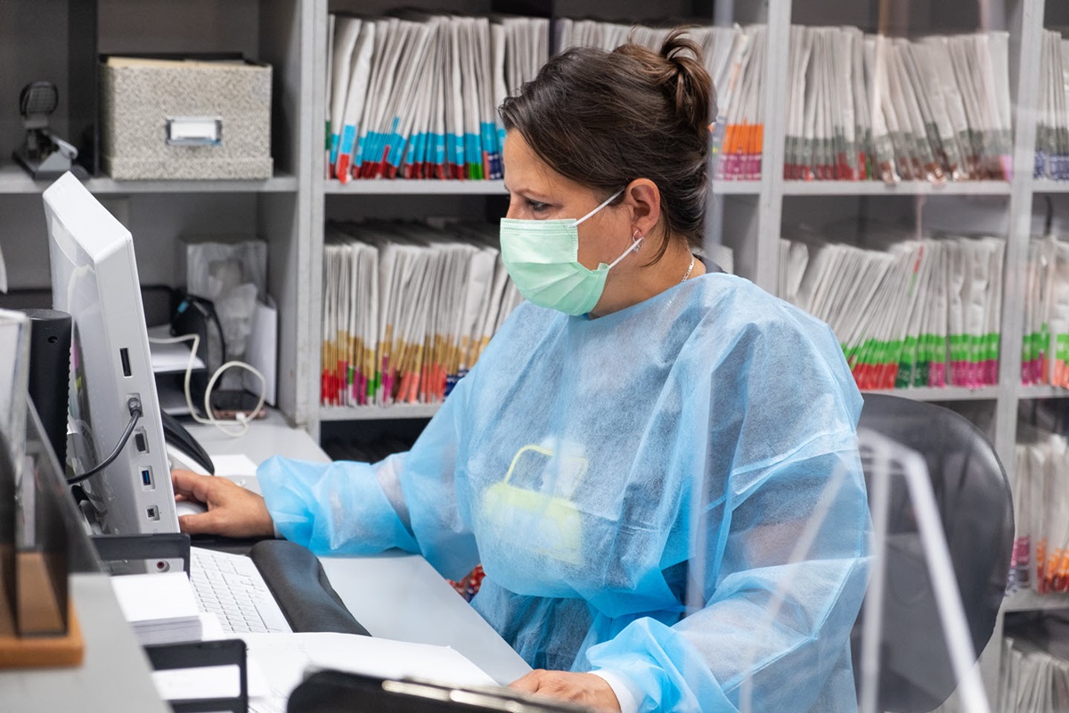 A woman wearing a surgical mask and gown sits at a desk looking at a computer, with medical files in the background.