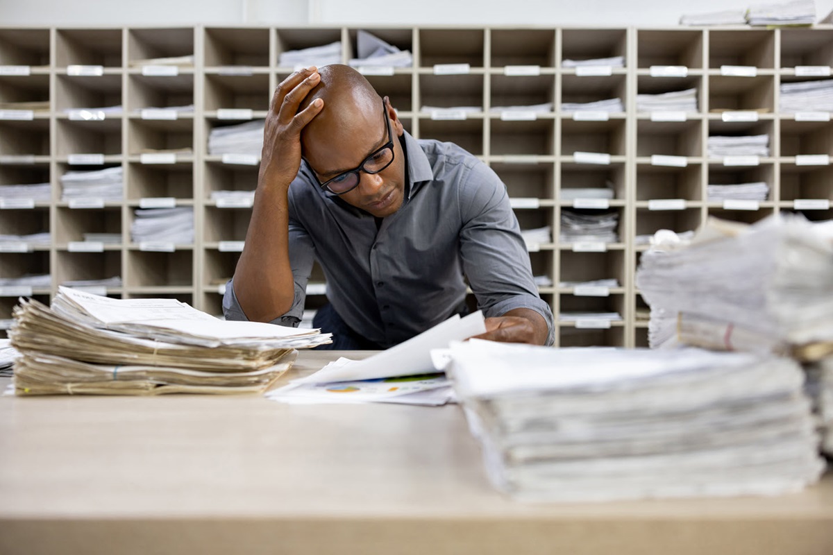 A man sits working at a table covered with paper documents. Behind him stand several filing cabinets full of paper.