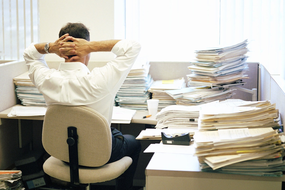 Man sitting in an office chair facing away from the camera and looking at a desk piled with documents. He appears overwhelmed and exhausted by the lack of organization.