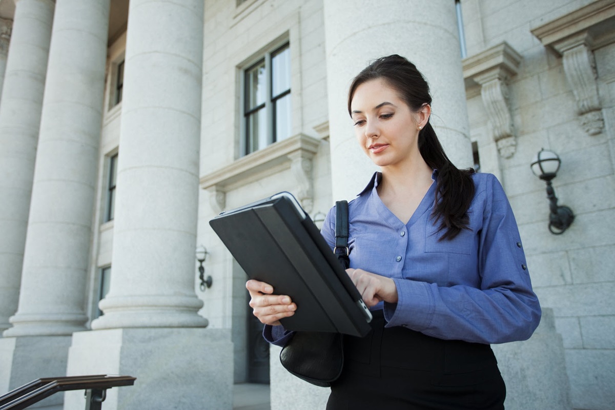 A woman with brown hair and a blue blouse stands on the steps in front of a building with large stone pillars. She is reviewing something on a tablet.