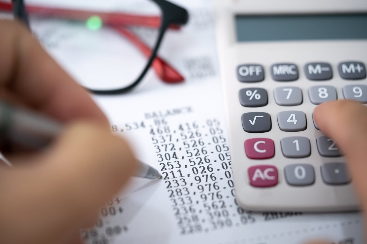 A person's hands use a pen to examine a balance sheet and operate a calculator.