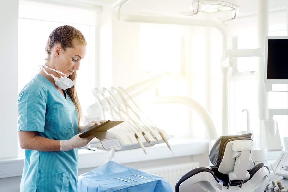 Side view of doctor using digital tablet. Professional is wearing scrub. She is standing by dentist's chair.