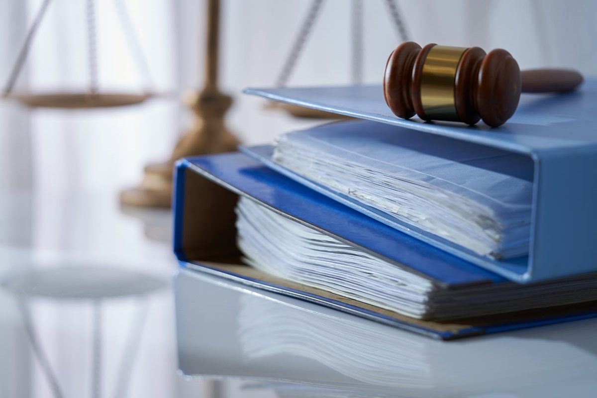 A gavel rests on a stack of two file folders filled with documents. A gold scale sits behind them.