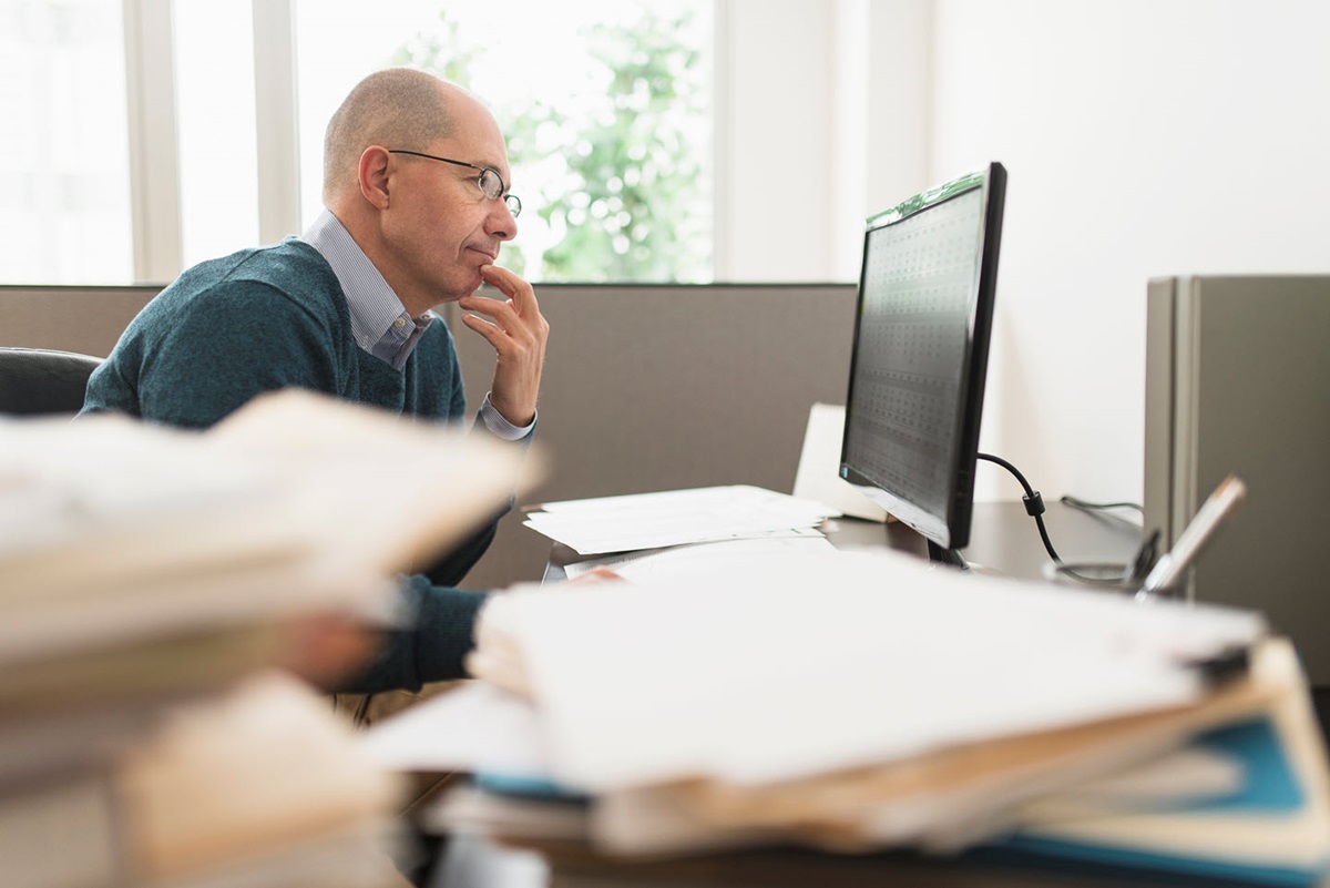 Stacks of out-of-focus file folders obscure the foreground. Behind them, a man works at a desktop computer.