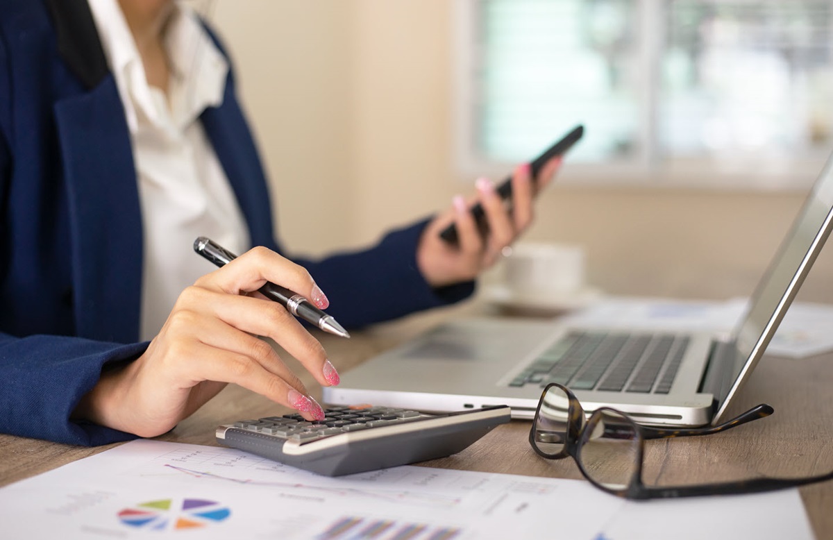 A businesswoman uses a calculator with one hand and a smartphone in the other. Her glasses, laptop, and business papers sit on the table in front of her.