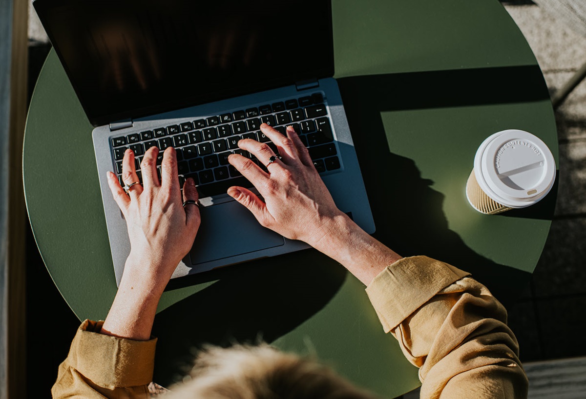 Top-down view of a laptop on a round green table, with a partially obscured person's two hands typing on it.