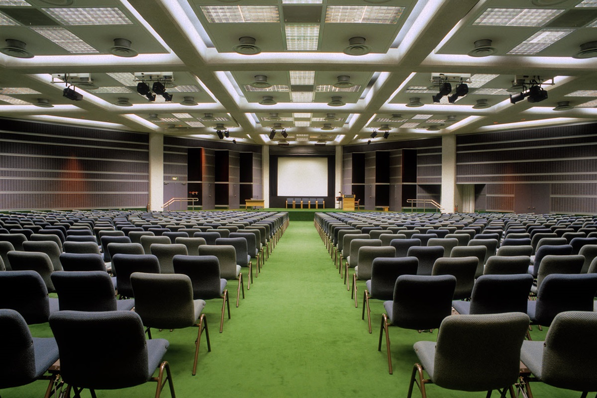 Rows of chairs are organized in a meeting hall with a clear aisle down the middle. Ahead on the stage, a projector screen is on display.