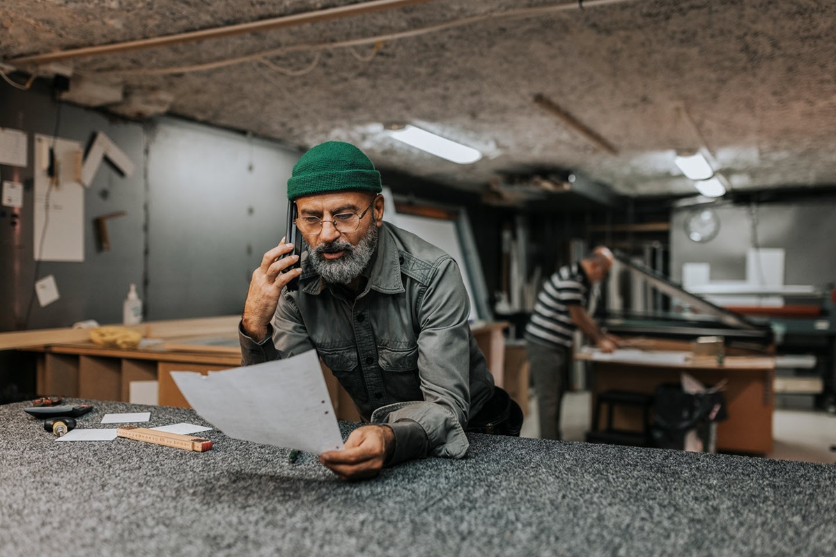 A small business owner leans on his workbench, talking on his phone with a customer while looking at a sheet of paper.