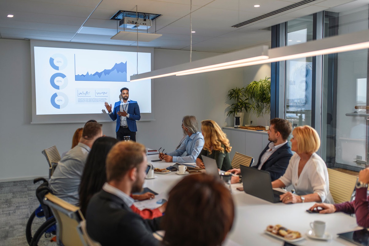 A man stands at the head of a business meeting, presenting charts and graphs displayed on a projector screen behind him.