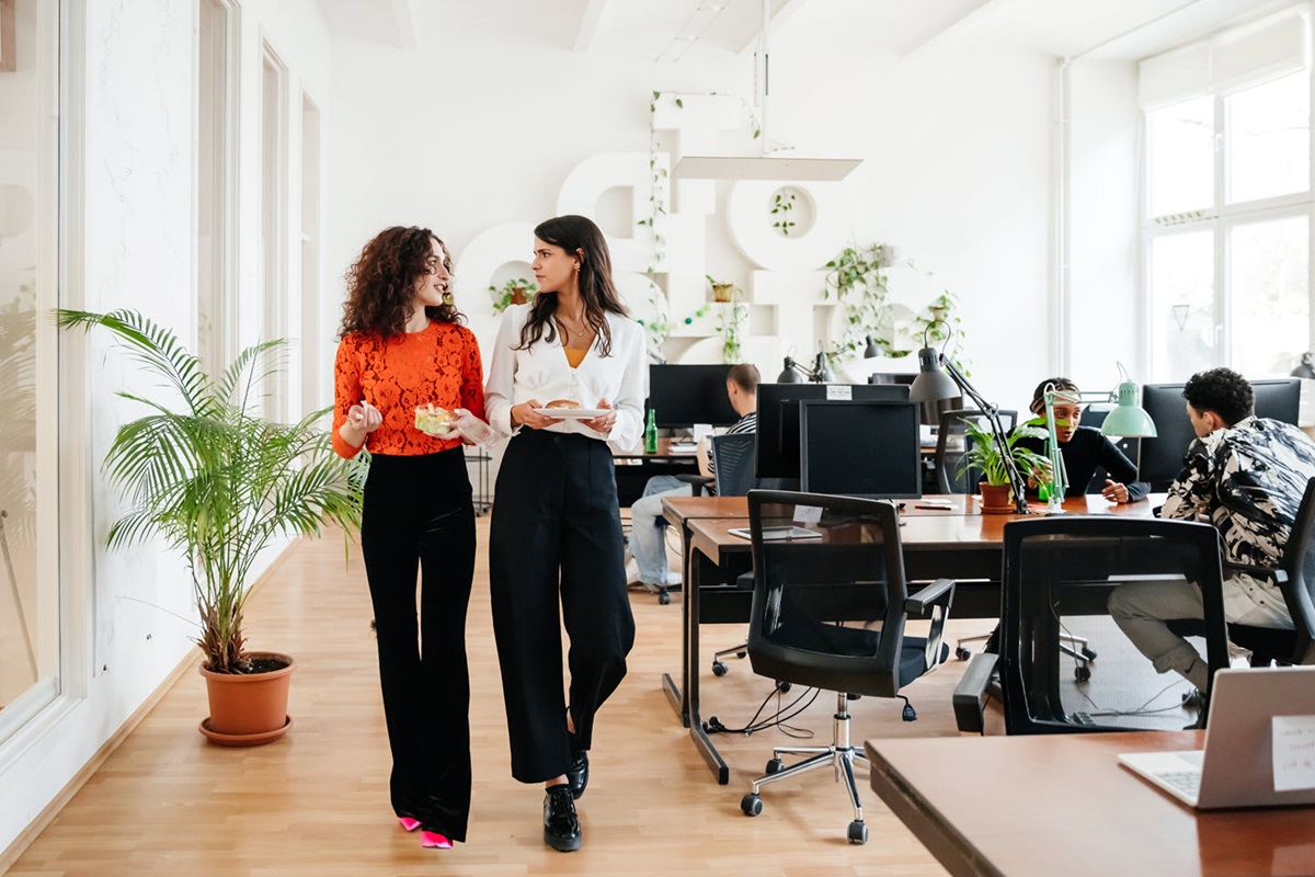 Two colleagues chat in a modern office filled with plants and natural light.