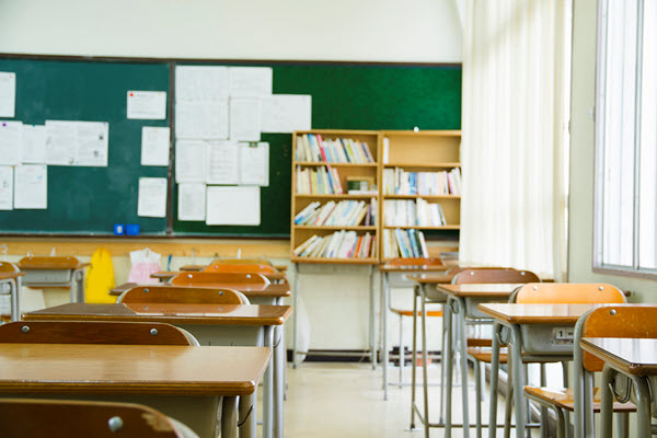 Desks inside a classroom with a chalkboard with papers in the backround and a bookcase.
