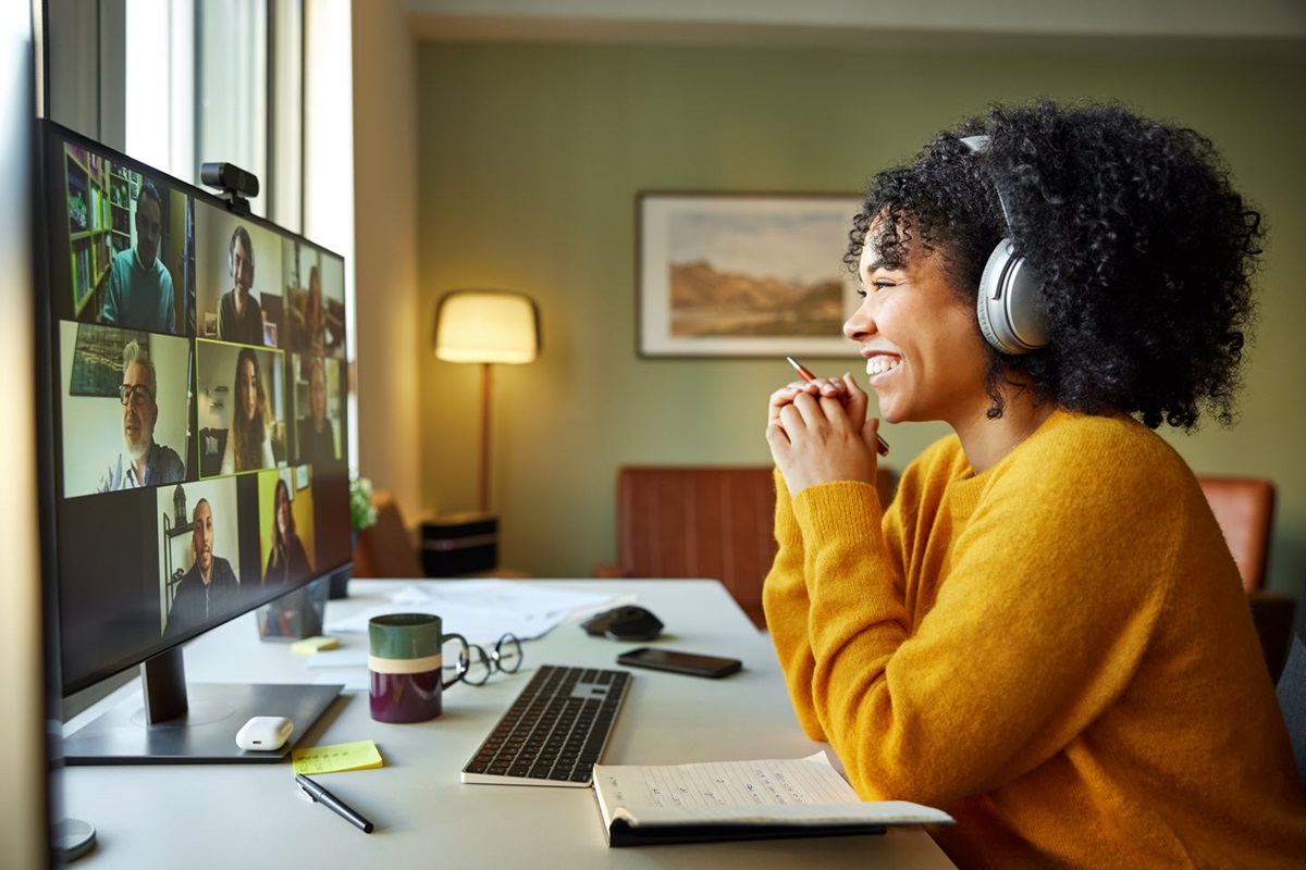 A woman sits at her desk in front of a computer screen. She's using video conferencing technology to sit in a meeting with several other coworkers.