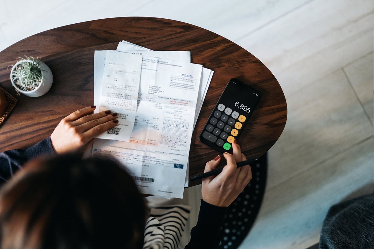 A woman sorts receipts and paperwork on the table in front of her. She's using her phone as a calculator.