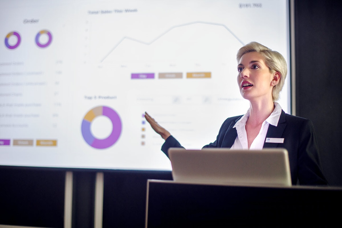 A businesswoman gives a presentation with a collection of graphs and charts projected on a screen behind her.