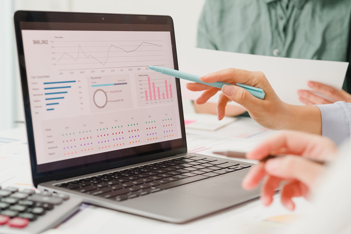 A hand holding a pen points to a chart on a laptop screen.