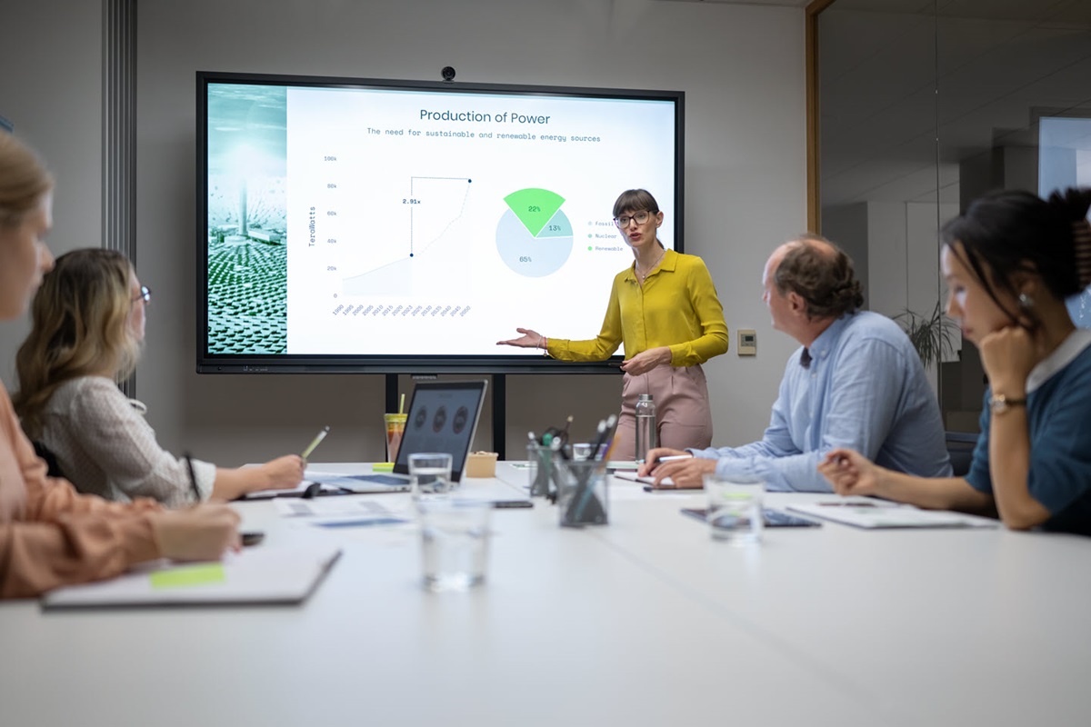 A gathering of workers looking at a projected image of a graph in a conference room with a standing woman pointing at the screen. 