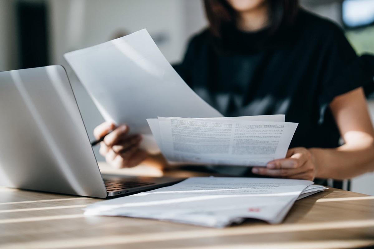The foreground shows a desk covered in paperwork and a laptop. A woman sits at the desk and holds several papers in her hands.