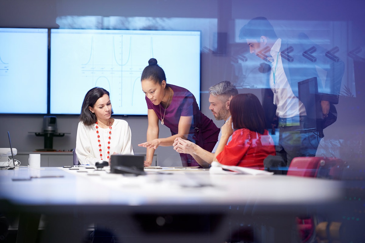 Several office workers in a glass conference congregate around a table. A screen behind them displays various graphs.