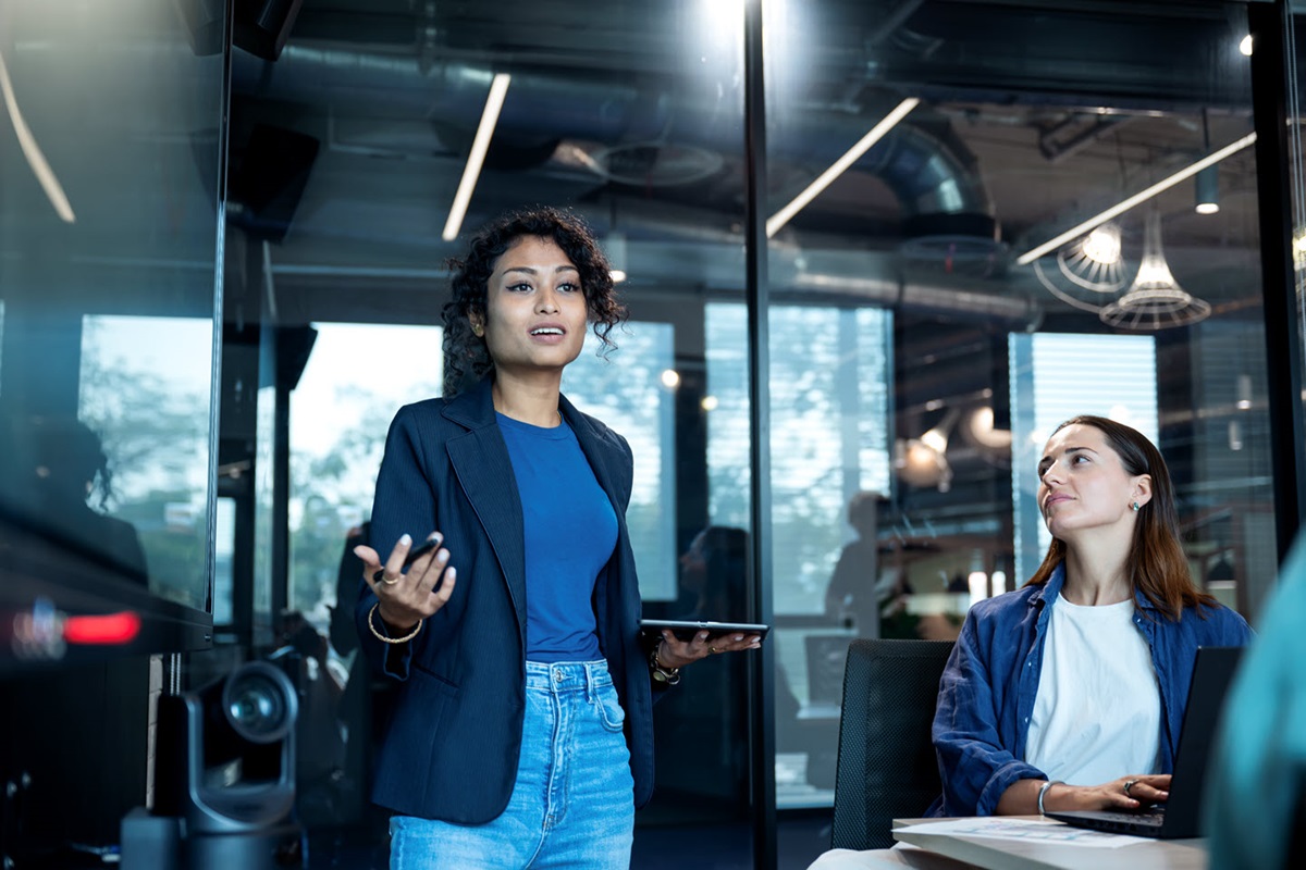 A consultant explains her plan for the company's digital transformation strategy in a meeting room. A woman sits to her left at the meeting room table to take notes on a laptop.