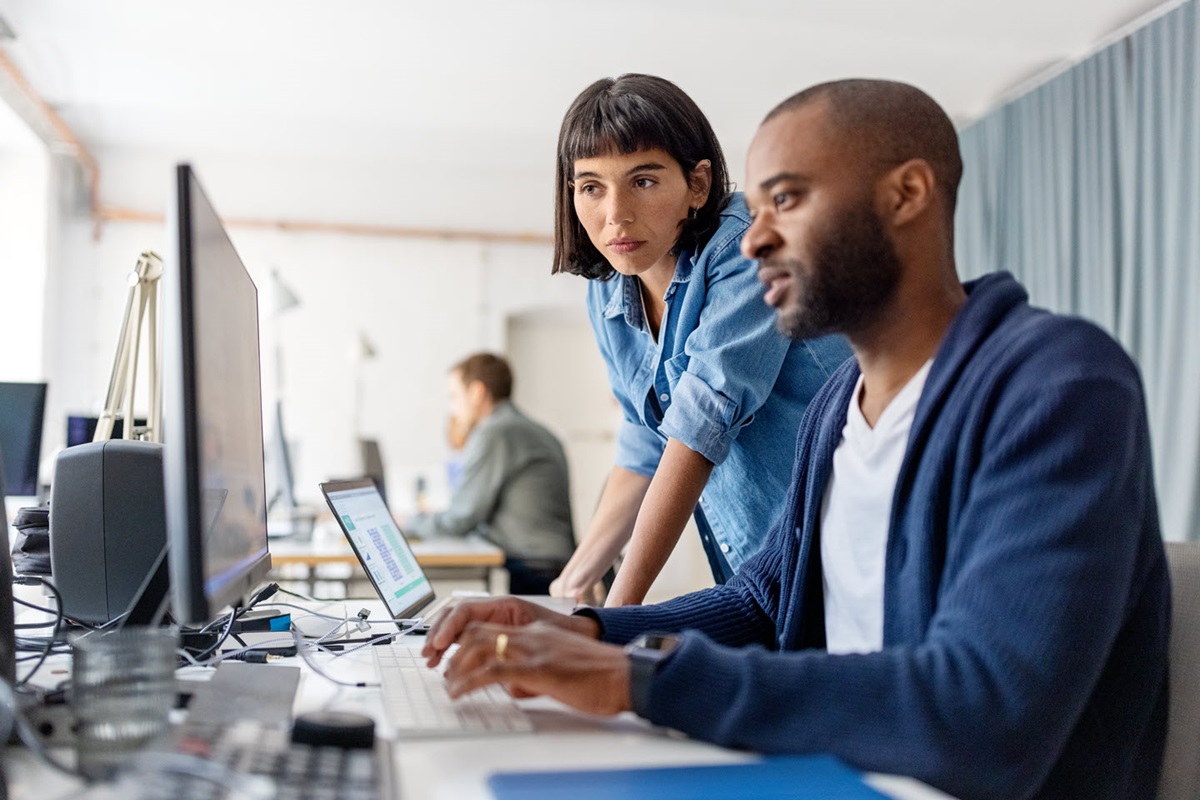 An employee sits at his desk typing on the keyboard as a co-worker looks over his shoulder to help.