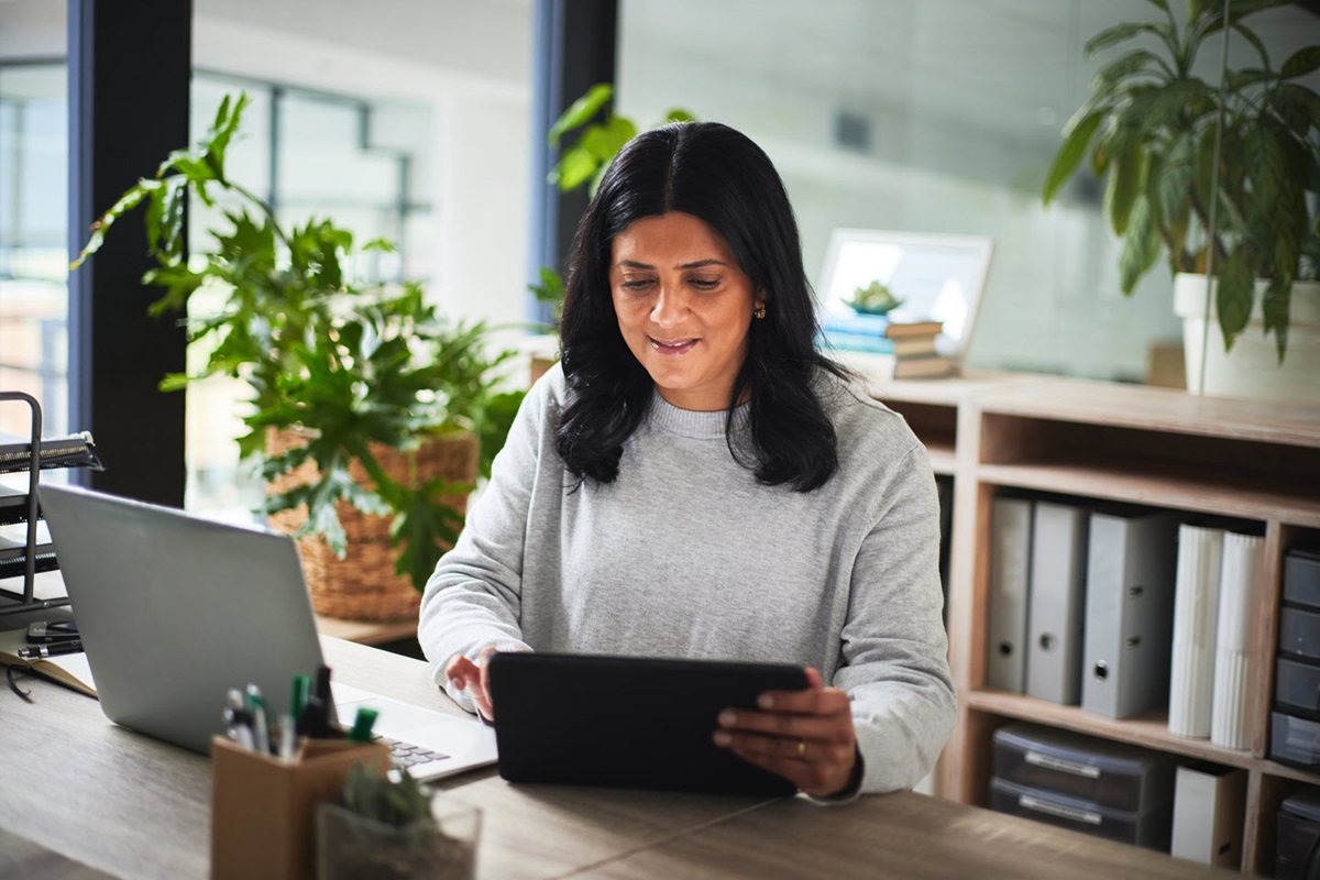 A woman works on a digital tablet at a wooden desk. There is a laptop in front of her and a shelf of binders behind her.