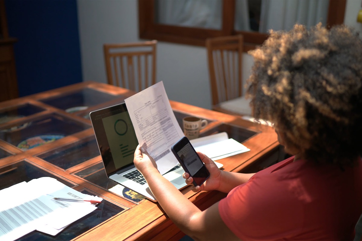 A woman sits at a dining room table with a laptop and some papers in front of her. She's taking a photo of a form with her phone.