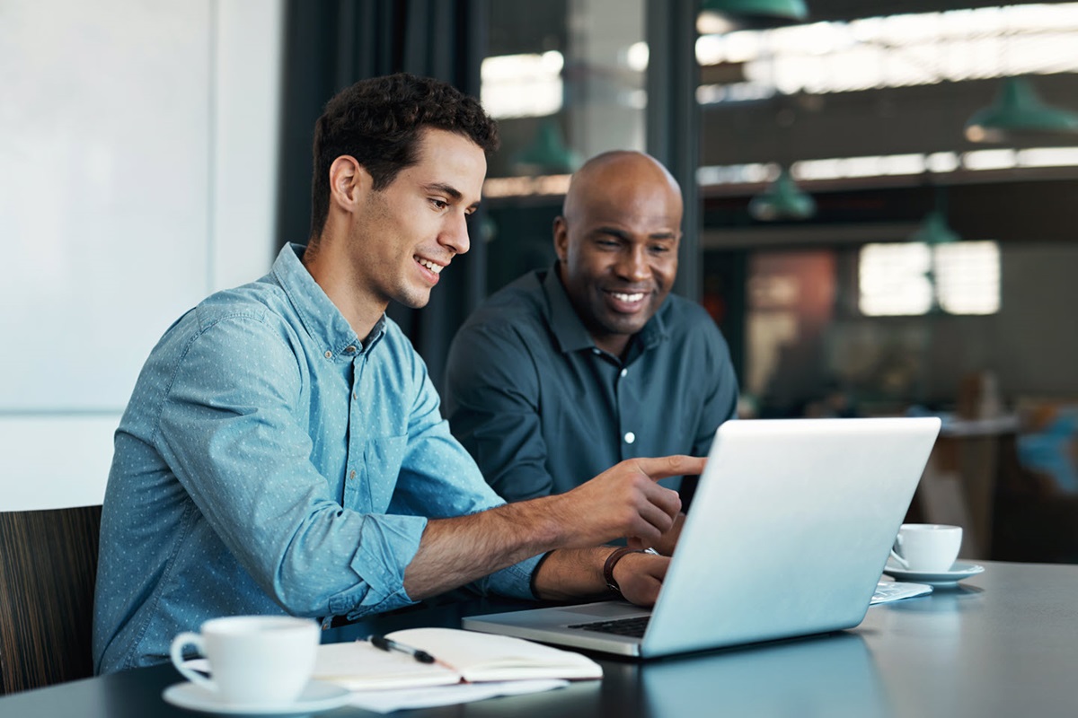 Two men work on a laptop in front of a glass window.