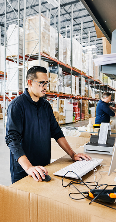 A man in a warehouse standing at a desk in front of a keyboard and monitor. 