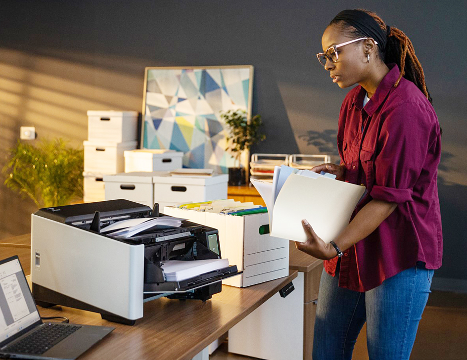 A woman standing next to a desk with a Ricoh fi-8950 document scanner holding a stack of papers.