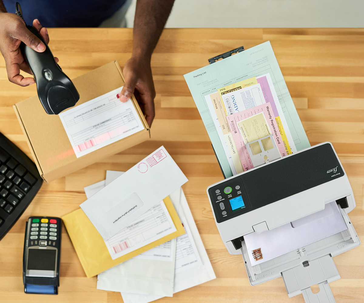 Overhead image of Ricoh scanner with work papers with a person holding a box and scanner int the other hand. 