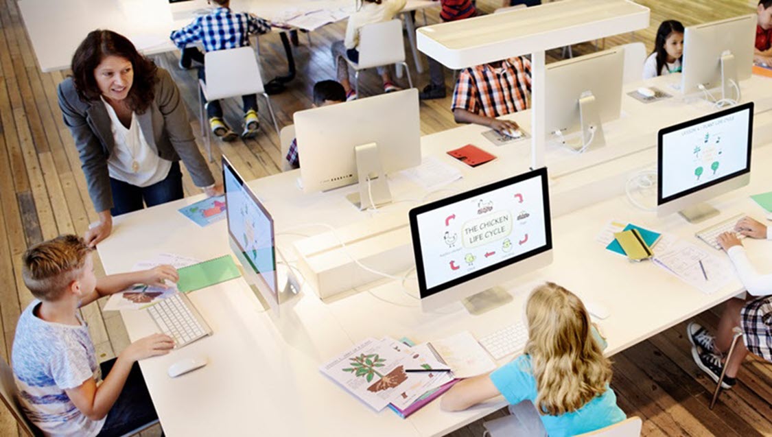 Teacher standing near students seated at computer desks. 