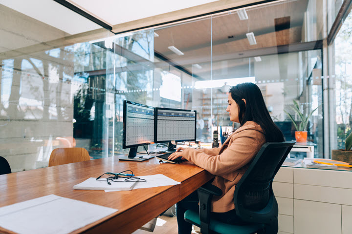 Side view of a Hispanic woman sitting at desk with two monitors. 