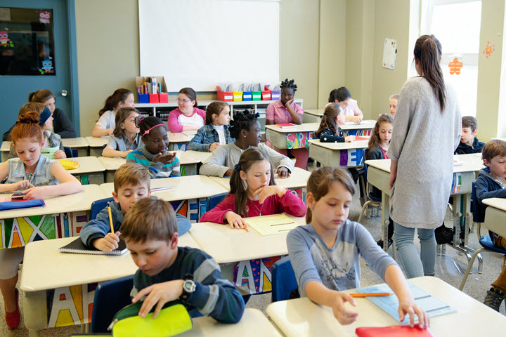 Classroom with children sitting at desks doing work while a teacher walks around. 