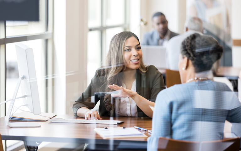 Hispanic executive woman behind a desk speaking with a female client. 