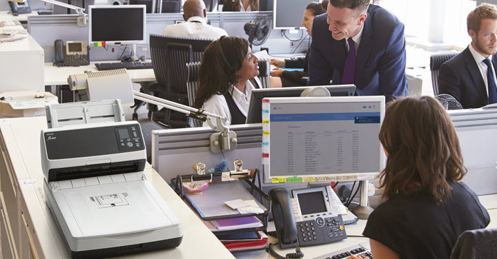 Image of the back of a woman sitting at a workstation in a busy office with other workers talking nearby and a RICOH fi-8270 on a shelf. 
