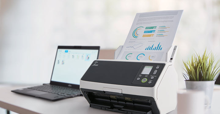 RICOH fi-8170 scanner with papers inside next to a laptop and some plants on a desk. 