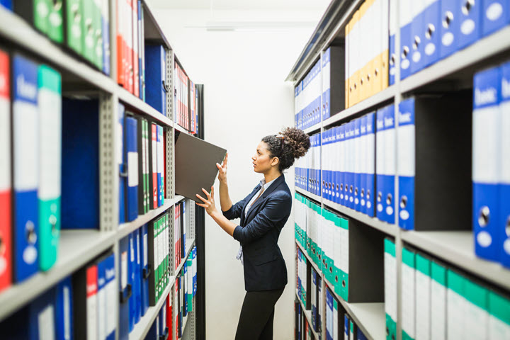 A woman, surrounded by neatly organized rows of binders, picks one up off of a shelf.