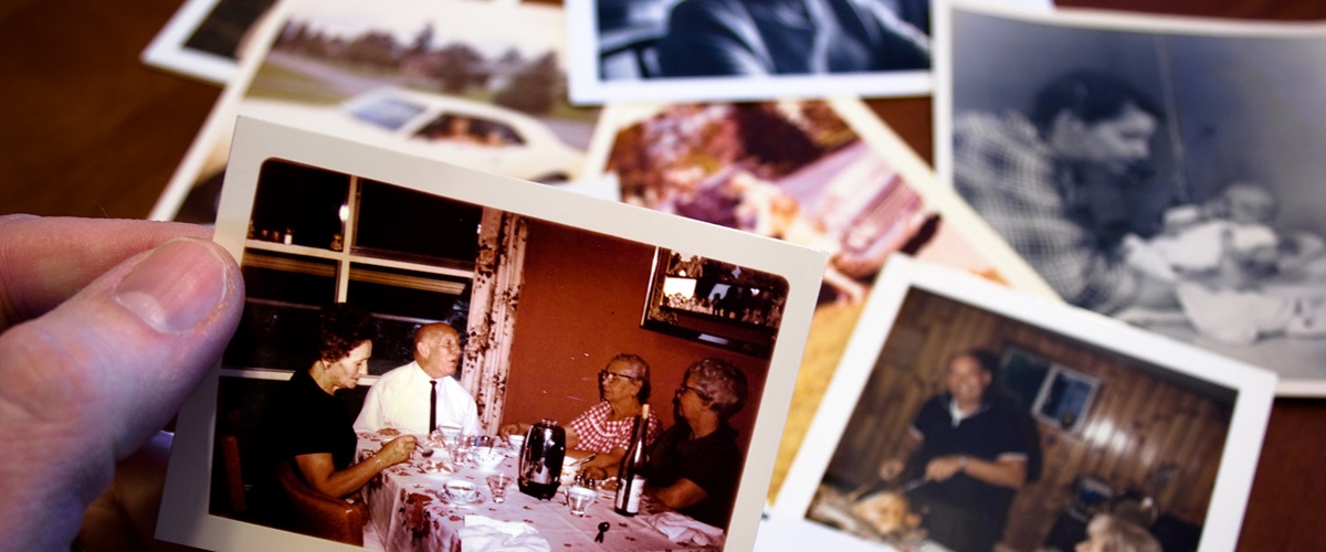 A hand holds up a small film print of four elderly people seated around a table wearing mid-century clothing. Behind that print, several more lie on a wooden surface.