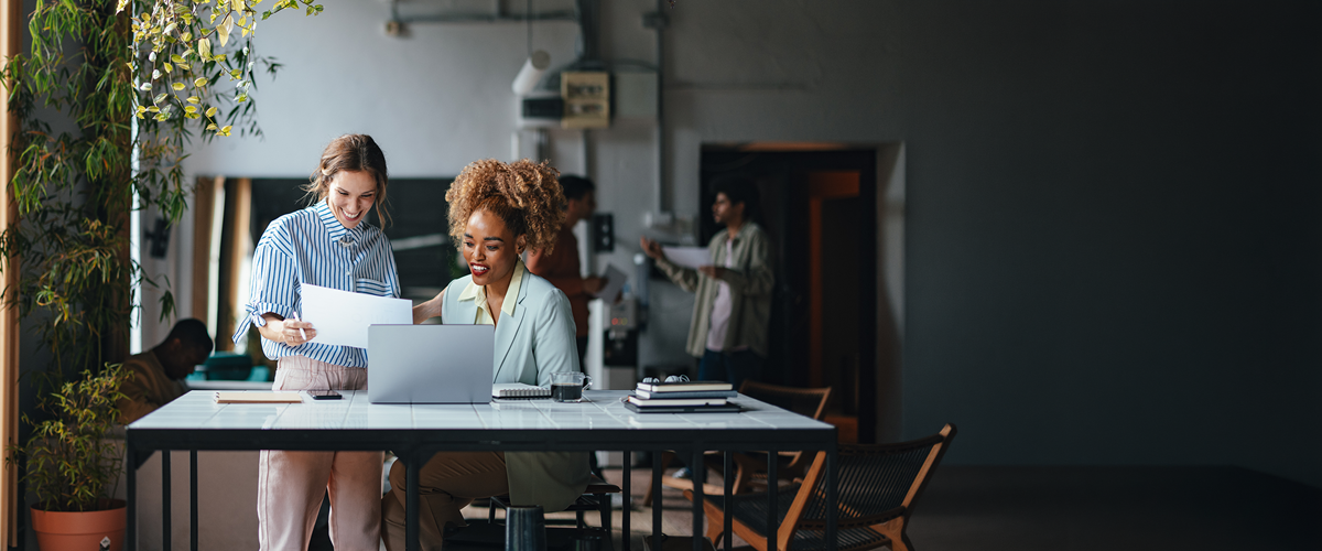 Two women in an office review paperwork in front of a laptop.