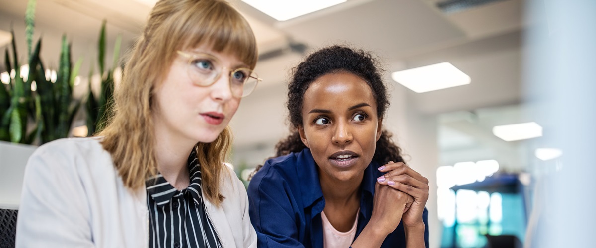 Two office workers sit at a desk and look at a computer monitor.