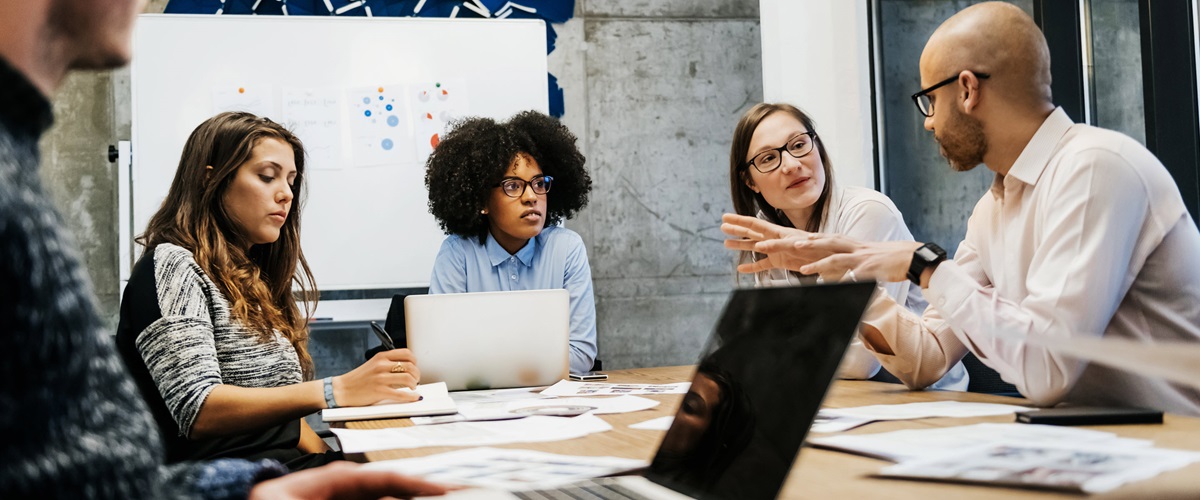 Three women and two men in a business meeting with laptops and paper in front of them.