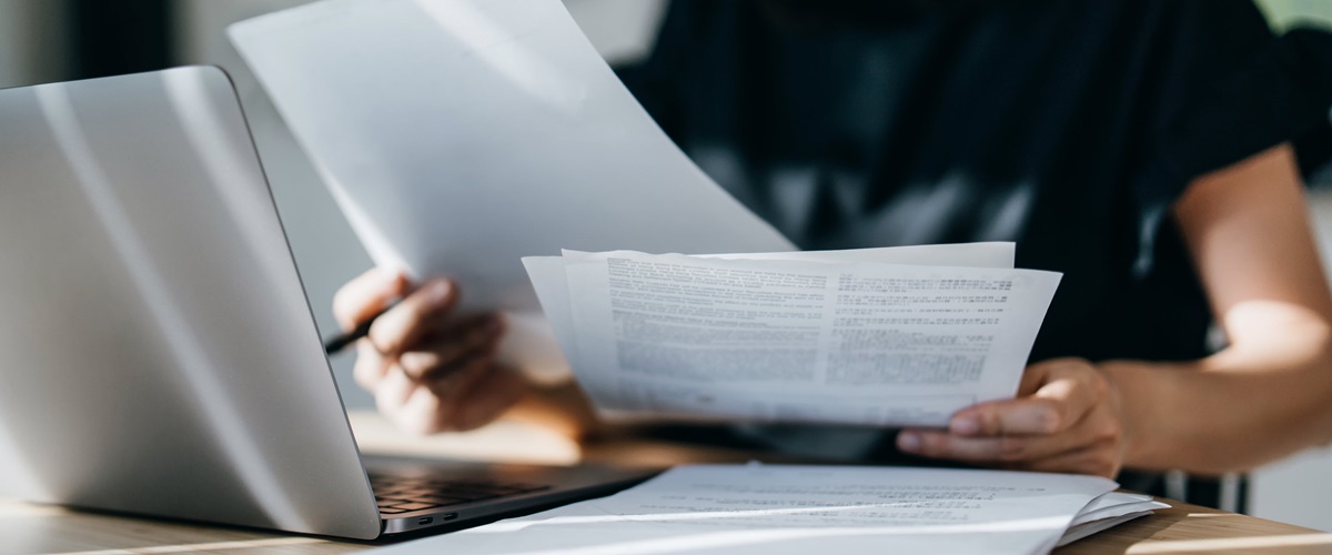 A person leafs through a small stack of papers while seated in front of a laptop.