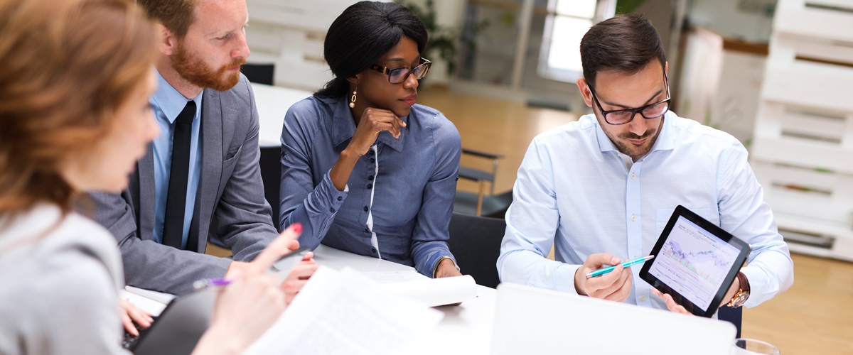 An office worker shows three colleagues information on a tablet while one refers to a paper document.