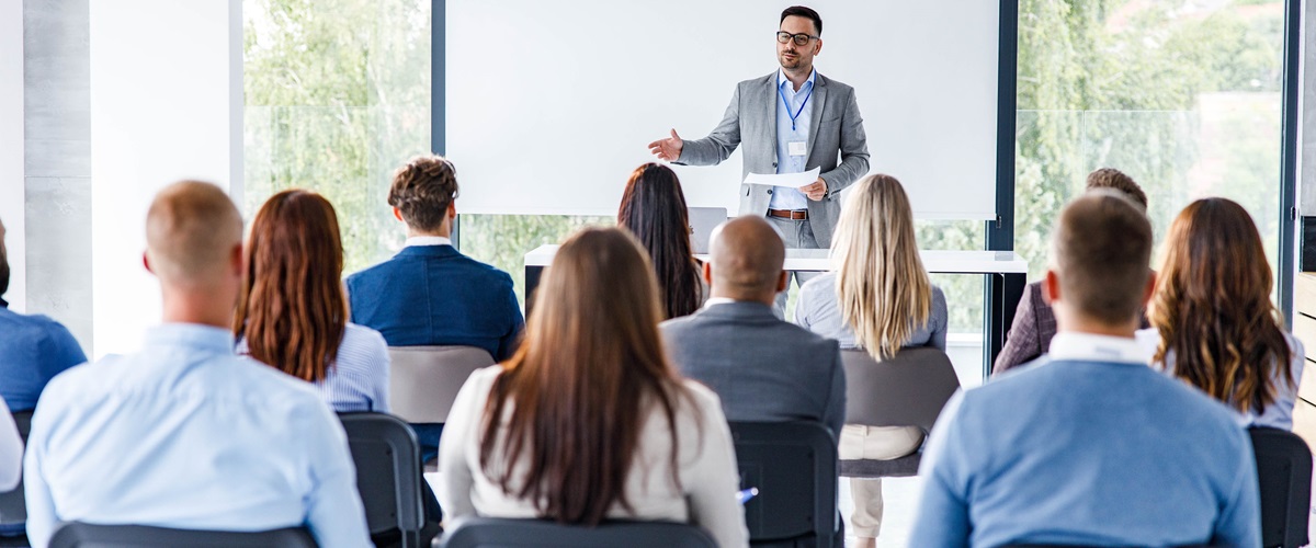 A man in a suit gives a presentation in front of a canvas screen for a group of businesspeople.